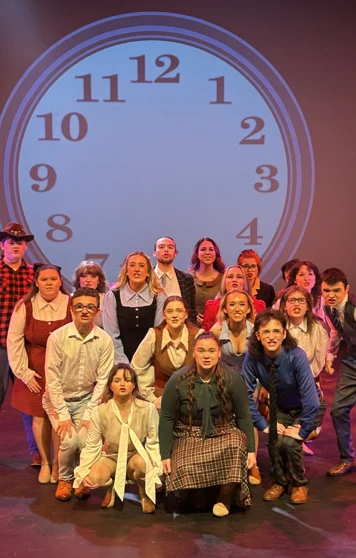 Group of theatre students posing for a cast photo in front of a large projected clock backdrop. Group of theatre students posing for a cast photo in front of a large projected clock backdrop.